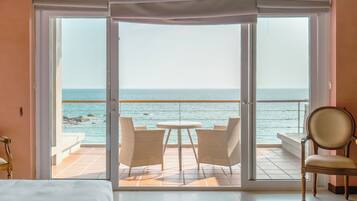 A hotel room with a large sliding glass door leading to a balcony with a view of the sea. There is a bed, a chair, and a small table on the balcony.