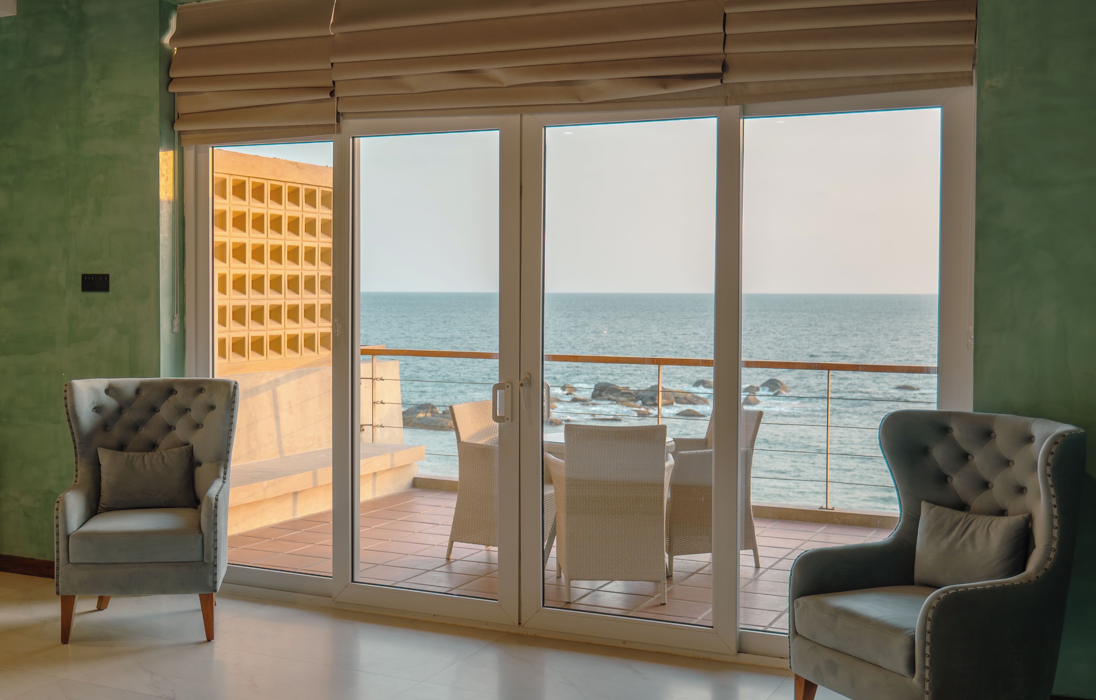 Ein Zimmer mit zwei Polstersesseln, einer Glastür, die auf einen Balkon mit Meerblick führt, und einem gefliesten Boden.