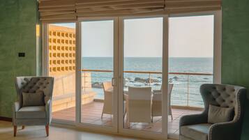 Ein Zimmer mit zwei Polstersesseln, einer Glastür, die auf einen Balkon mit Meerblick führt, und einem gefliesten Boden.