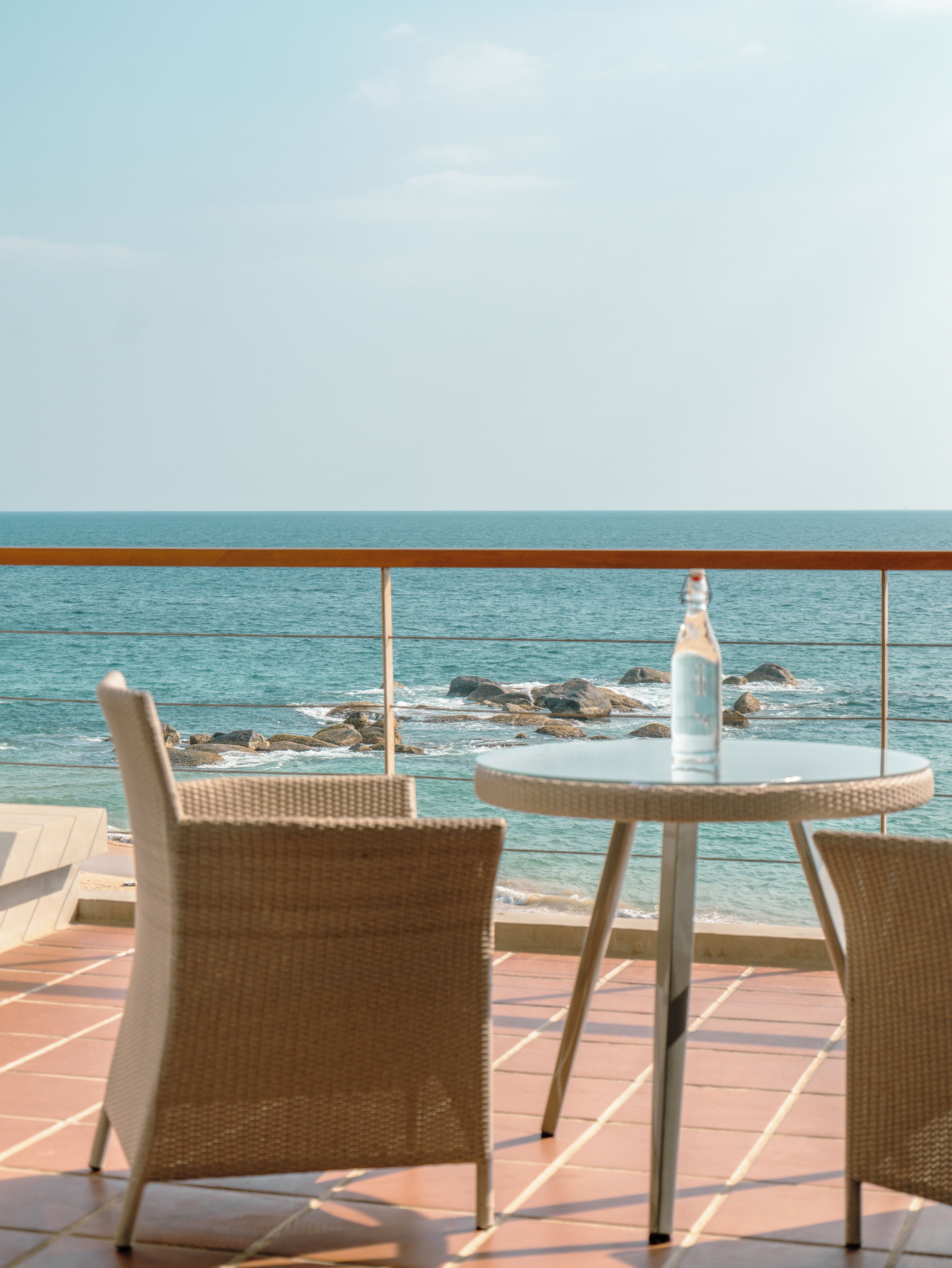 Een ronde tafel met twee rieten stoelen op een terras met uitzicht op zee, met een fles op de tafel.