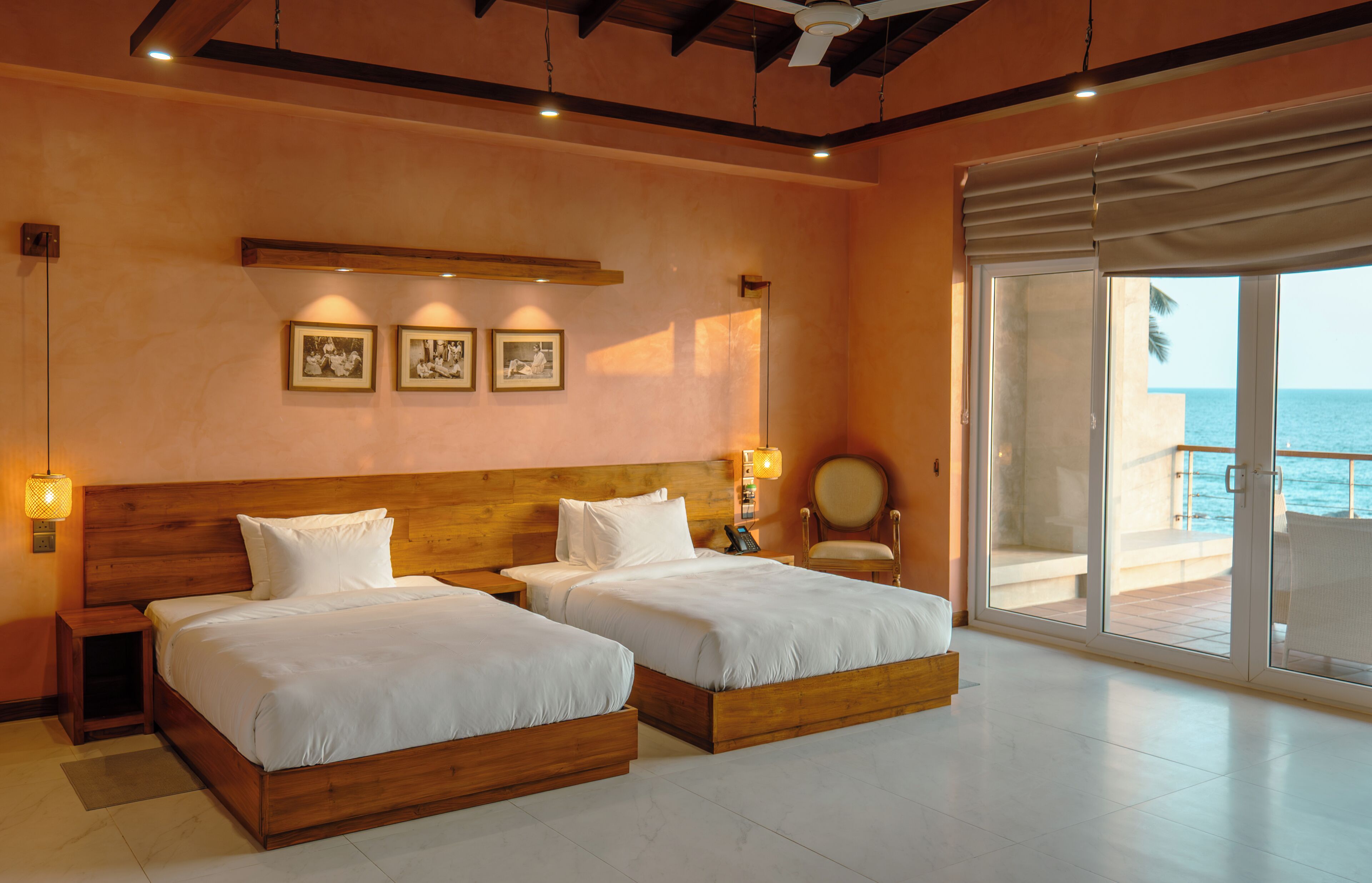 A hotel room with two beds, a wooden headboard, a large window with a view of the sea, and a wall-mounted light fixture.