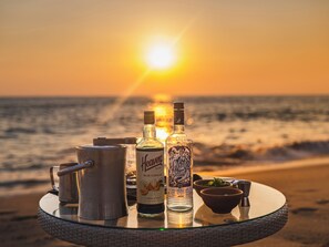 Une table avec une bouteille de spiritueux, un pichet en métal et un bol, sur une plage au coucher du soleil.