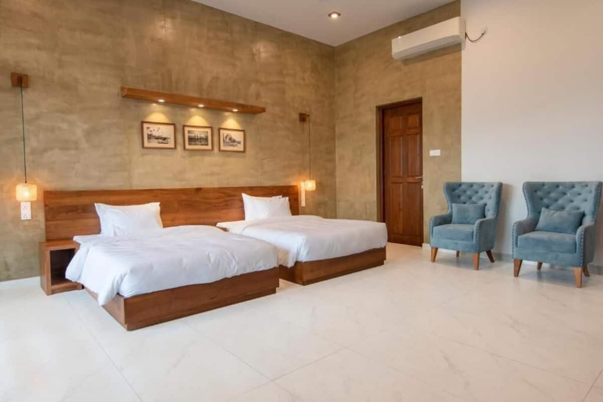 A bedroom with two beds, a wooden headboard, a wall-mounted shelf with pictures, and two upholstered chairs.