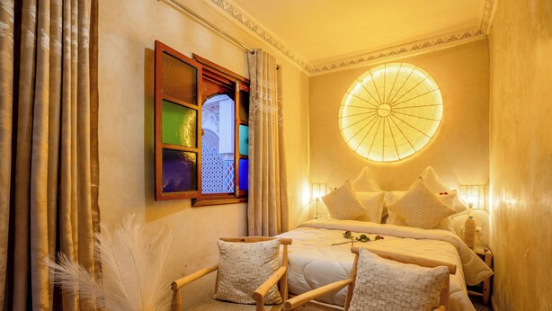 A cozy bedroom with a bed, two chairs, a window with curtains, and a wall-mounted light fixture. - Riad le Graff Pink (Marrakech)
