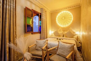 A cozy bedroom with a bed, two chairs, a window with curtains, and a wall-mounted light fixture. - Riad le Graff Pink (Marrakech)
