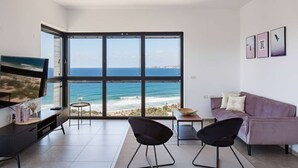 A modern living room with a large window overlooking the ocean, a sofa, two chairs, a coffee table, and a TV on a stand. - Family Getaway Sea View Parking (Bat Yam)
