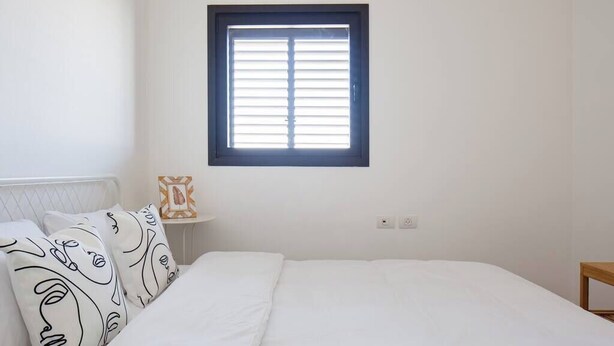 A bedroom with a bed, white bedding, a window with blinds, a bedside table with a lamp, and a wall-mounted power outlet. - Family Getaway Sea View Parking (Bat Yam)
