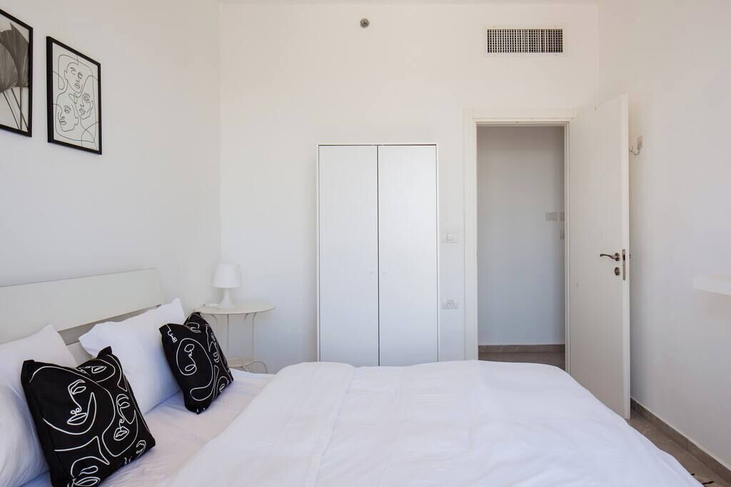 A bedroom with a bed, white walls, a wardrobe, and a door leading to another room.