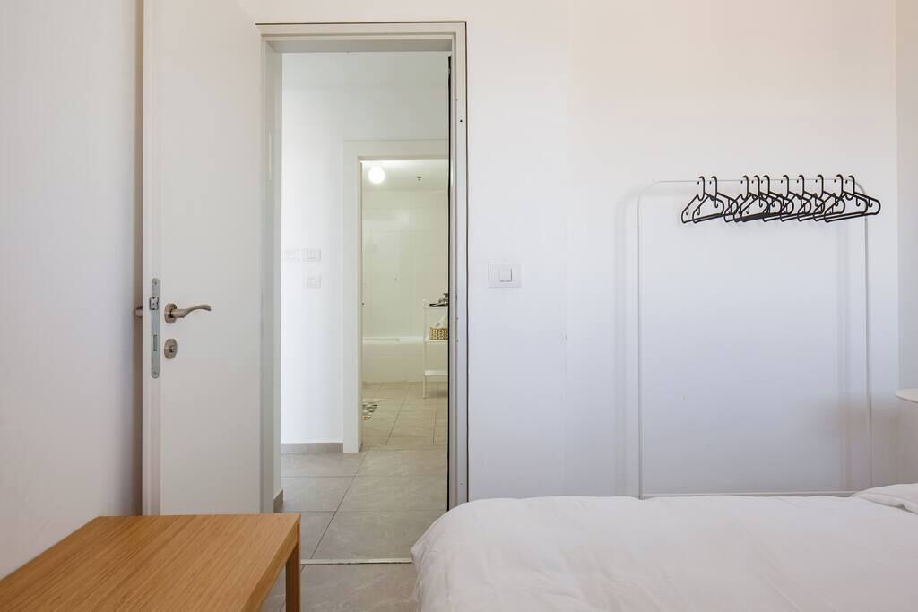 A hotel room with a wooden bench, a bed, a coat rack, and a door leading to a bathroom.