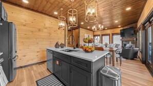 A modern kitchen with a central island, wooden walls, and a dining area with a TV and fireplace. - Rooftop Hot Tub, Indoor Pool, Sunrise & Mtn Views (Pigeon Forge)