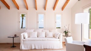 Ein Zimmer mit einem weißen Sofa, einem kleinen runden Tisch, einer Lampe und Meerblick durch die Fenster.