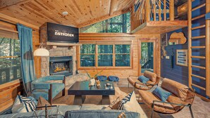 L’intérieur chaleureux d’un chalet avec une cheminée en pierre, un plafond en bois et de grandes fenêtres donnant sur la forêt.