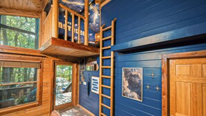Une cabane avec une chambre à coucher située en mezzanine, une échelle en bois et une grande fenêtre donnant sur une terrasse.