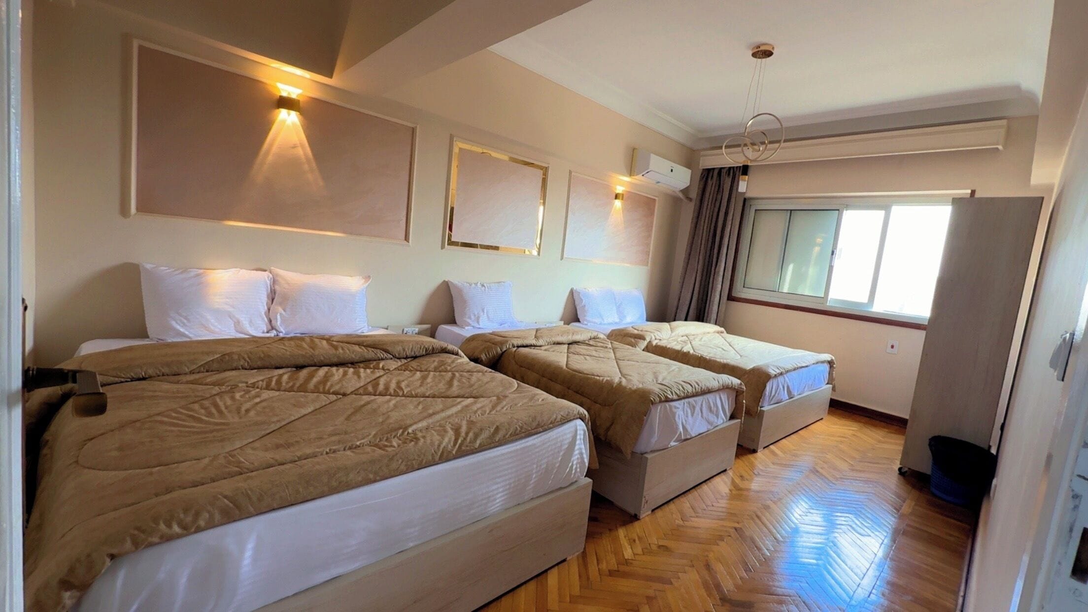 A hotel room with three beds, a wooden herringbone floor, and a window with curtains.
