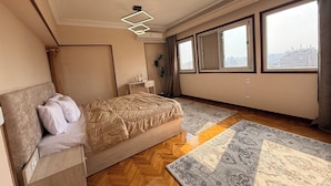 A bedroom with a bed, a nightstand, a dresser, a large window with a city view, and a modern ceiling light fixture. - Golden mile - Downtown Cairo (Cairo)