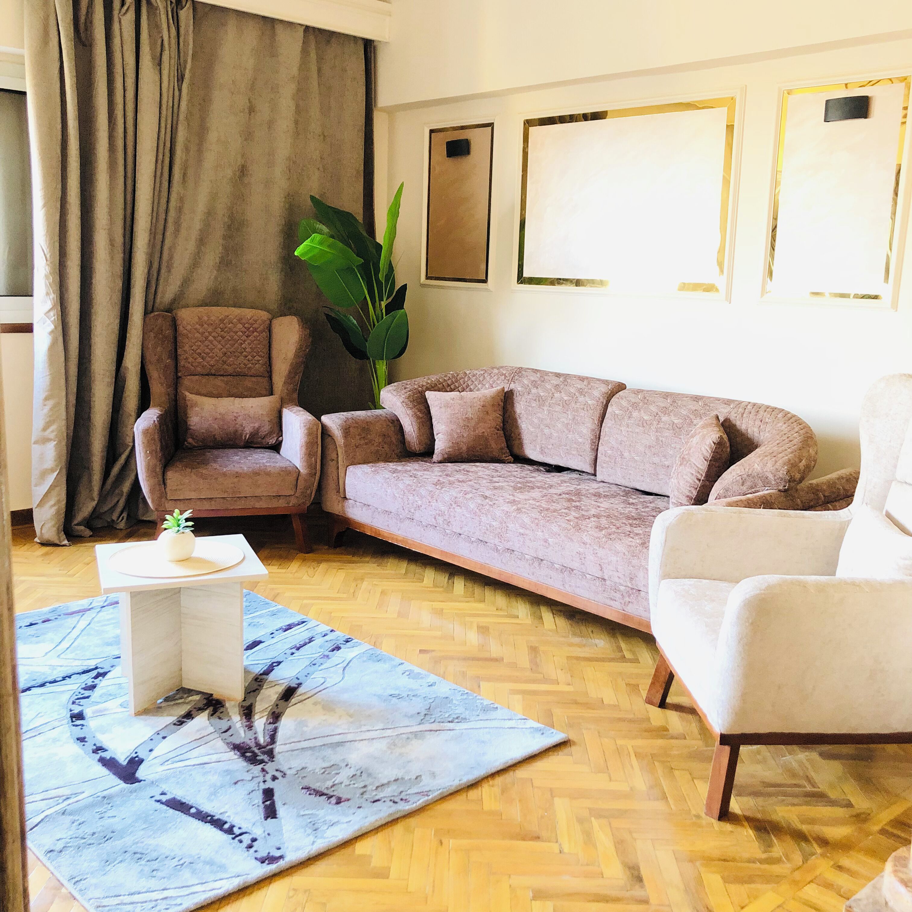 Ein Wohnzimmer mit braunem Sessel, einem beigen Sofa, einem weißen Sessel, einem Couchtisch und einer großen Grünpflanze.