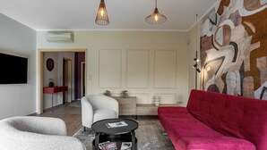Una sala de estar moderna con un sofá rojo, un sillón blanco y una mesa de centro negra. Hay un televisor de pantalla plana montado en la pared, una mesita auxiliar roja y una alfombra estampada.