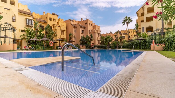 Una piscina con borde de baldosas, rodeada de palmeras y edificios con balcones.