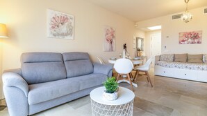 Una sala de estar moderna con un sofá gris, una mesita redonda blanca con una planta, un área de comedor con una mesa y sillas blancas, y un cuadro floral en la pared.