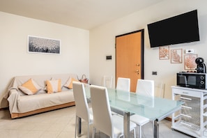 Un moderno salón con una mesa de vidrio, sillas blancas, un sofá con cojines, un televisor de pantalla plana y un microondas sobre un carrito blanco.