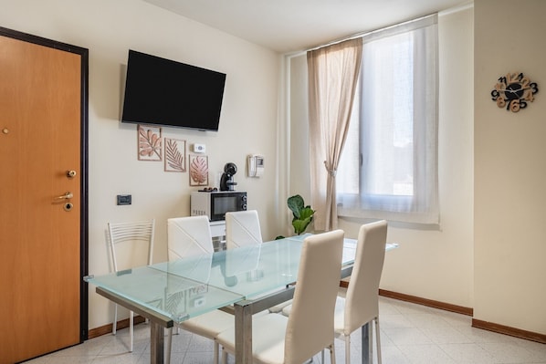 Un comedor con una mesa de vidrio, sillas blancas, un televisor en la pared, una ventana con cortinas y un reloj.