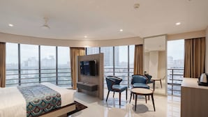 Una habitación de hotel moderna con una cama grande, un televisor, dos sillones, una mesita y vistas a la ciudad a través de amplios ventanales.