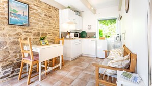 Une pièce chaleureuse avec un mur en pierre, une table à manger en bois avec des chaises, une petite cuisine blanche et un lit en bois avec une literie à motifs floraux.