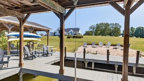 A wooden dock with chairs and tables, a covered area with umbrellas, and a view of a house and lawn.