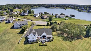 An aerial view of a residential area with houses, a large lake, and a prominent white house with a porch and a staircase leading to the entrance.