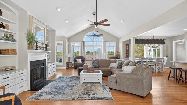 A spacious living room with a fireplace, a ceiling fan, and a large window offering a view of the outdoors.