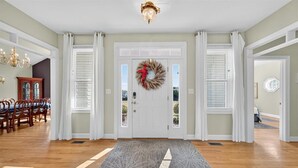 A spacious entryway with a wooden floor, a decorative rug, a white door with a wreath, and a dining area with wooden chairs and a table.