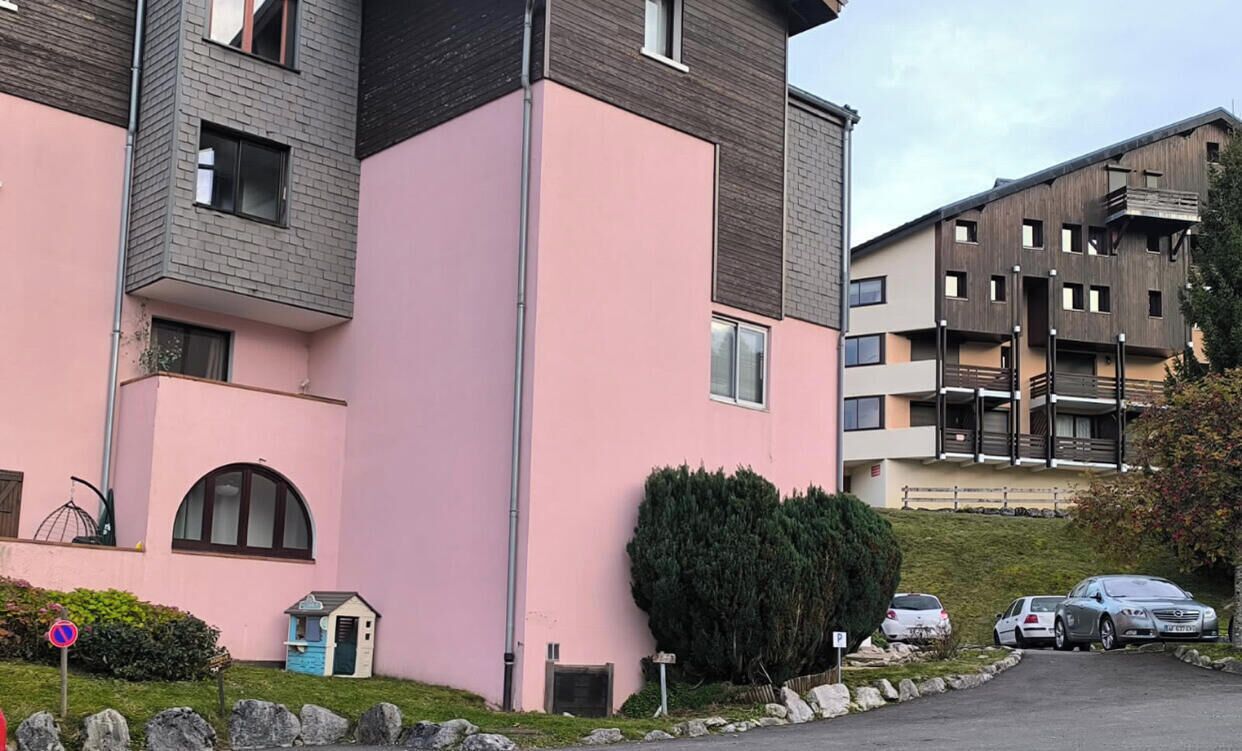 A multi-story residential building with a pink facade, a small garden area with a shed, and a parking lot with cars.