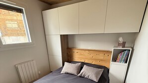 A bedroom with a bed, wooden headboard, white cabinets, a radiator, and a window with a view of a wooden building.