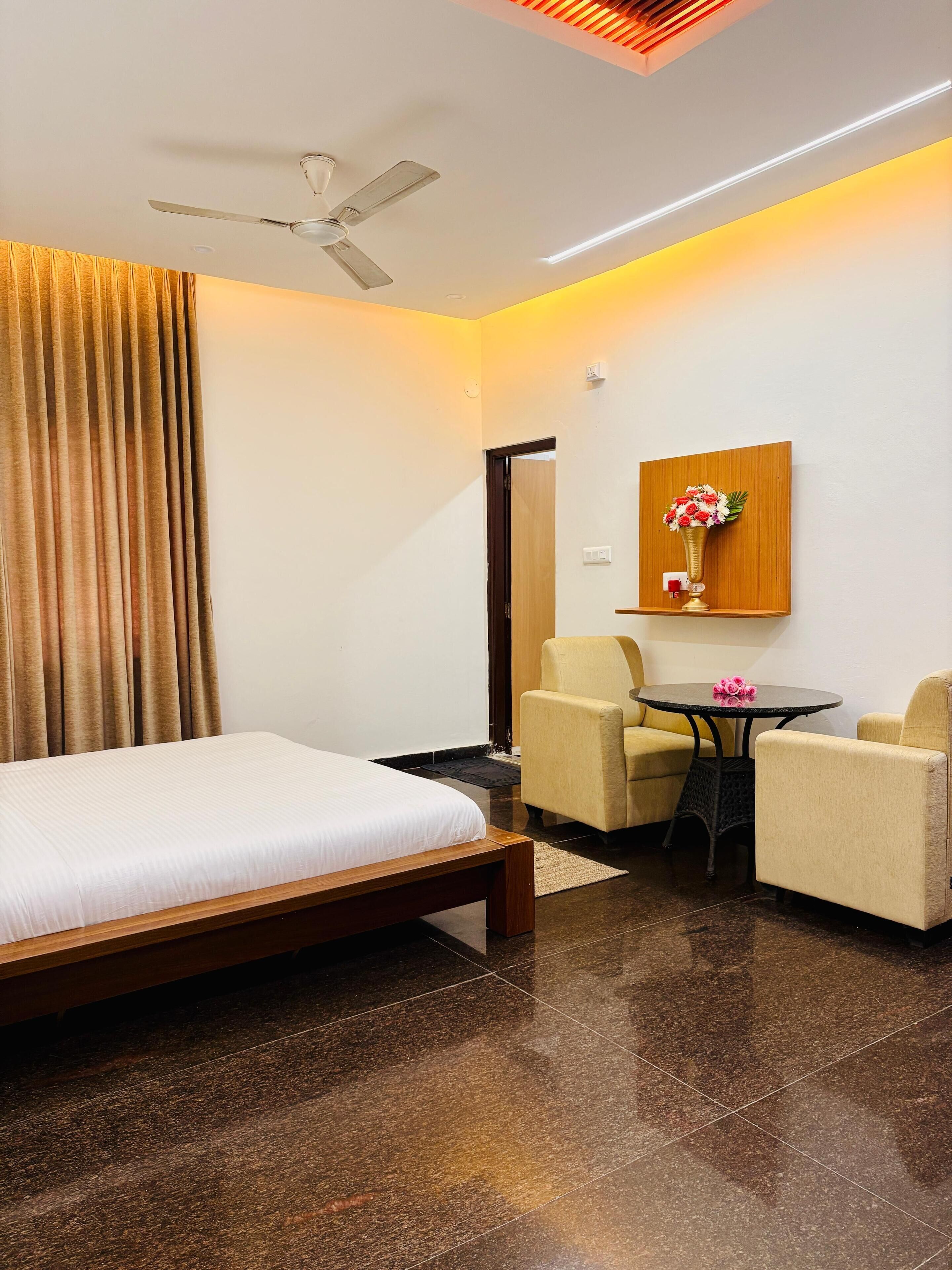 A hotel room with a bed, two armchairs, a small table, a ceiling fan, and a wall-mounted vase with flowers.