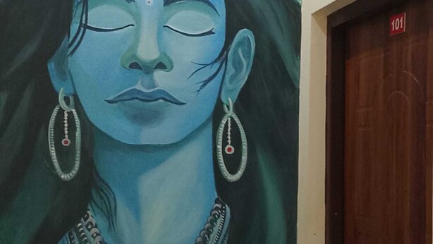 A mural of a figure with a serene expression, wearing traditional jewelry, in a hallway with a wooden door and a blue painted wall. - Mandakini River View (Jakholi)