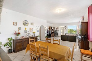 A dining area with a table and chairs, a kitchen in the background, and a view through a doorway.