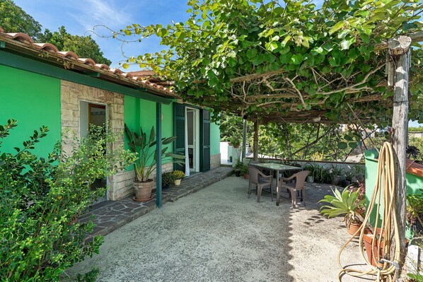 A green building with a stone entrance, a covered patio with a table and chairs, and various potted plants.