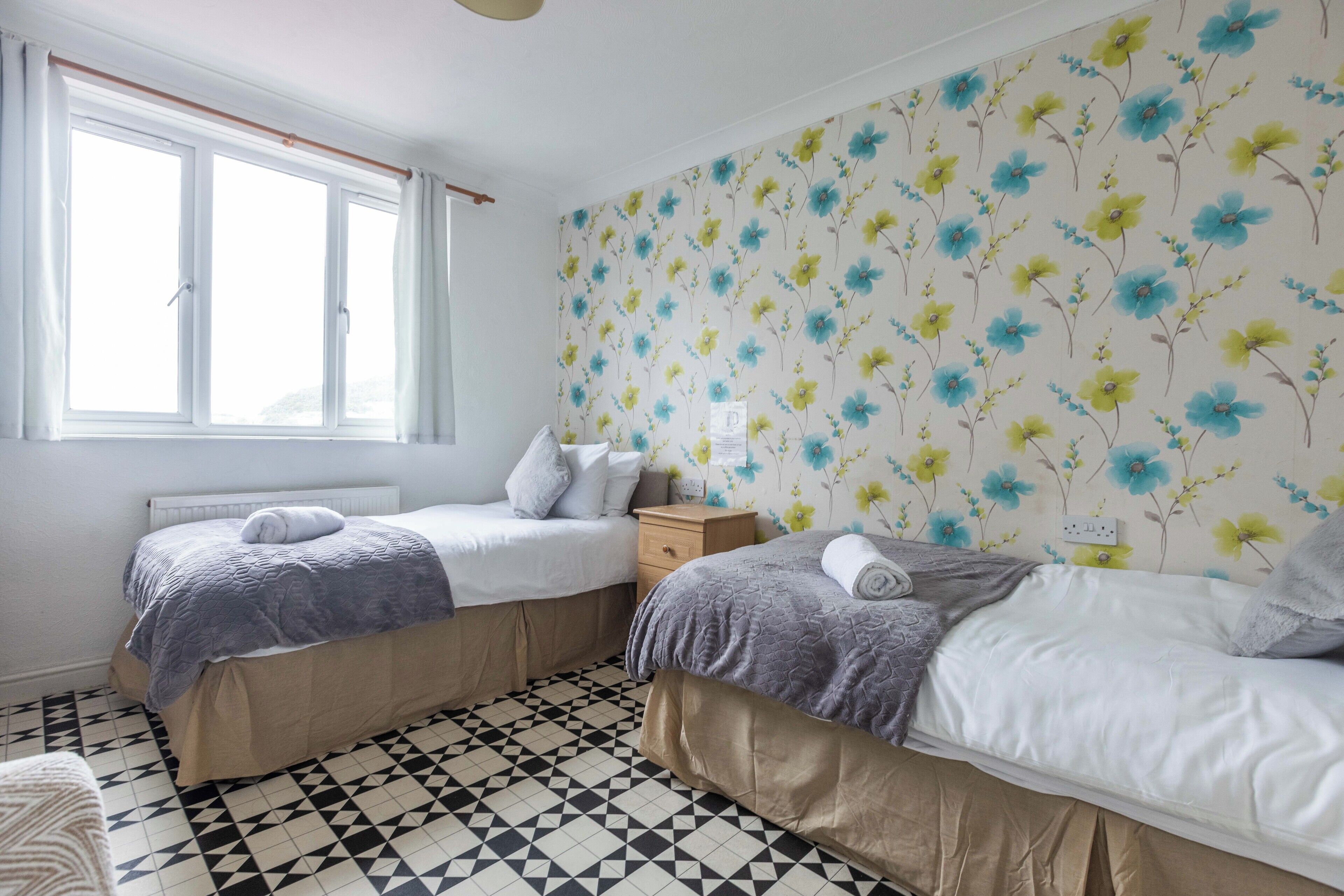 Una habitación con dos camas, una ventana, un radiador y un papel tapiz estampado.