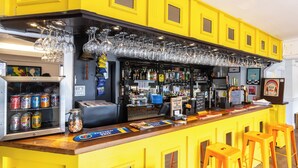 Zona de bar con decoración amarilla, que incluye una barra con taburetes, una vitrina de cristal y diversas botellas y latas.