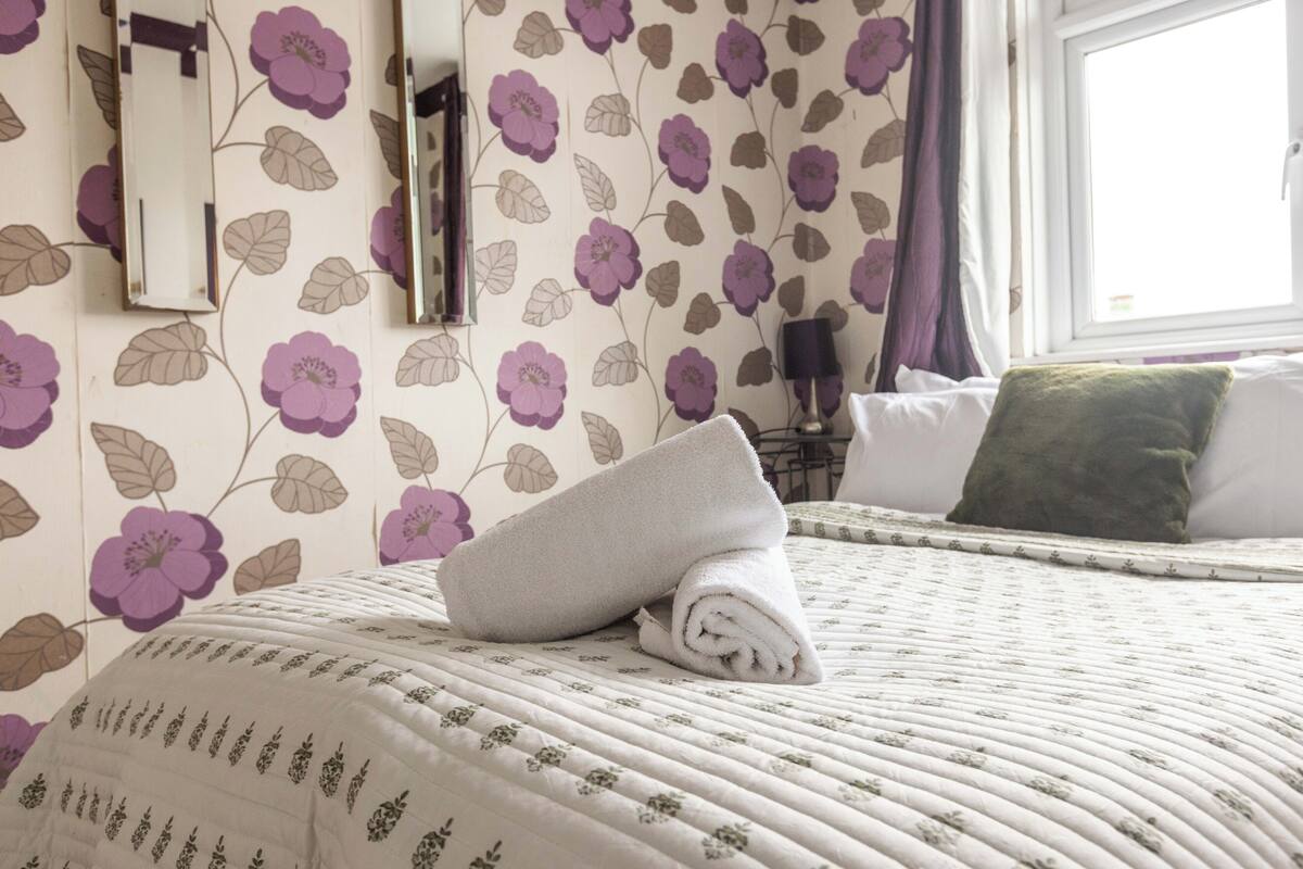 Een netjes opgemaakt bed met een opgerolde handdoek, een groen kussen en een wand met bloemenmotief.