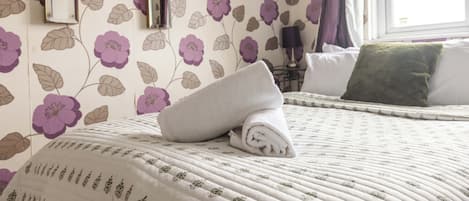 Una cama bien hecha con una toalla enrollada, una almohada verde y un papel tapiz estampado con motivos florales.