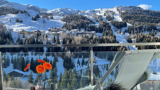 A balcony with a view of a snowy mountain landscape, a glass table with a vase of orange flowers, and a chair. - Apartment Bergjuwel (Hermagor)