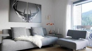 A modern living room with a grey sofa, a smaller grey armchair, a white furry throw, a wall-mounted deer artwork, a small table with a vase of flowers, and a view of a mountain through a large window. - Apartment Bergjuwel (Hermagor)
