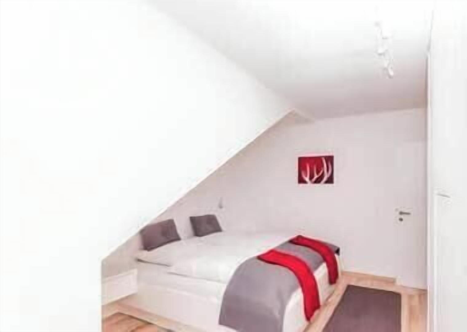 Habitació petita de color blanc amb teulada inclinada, un llit, una porta i un llum fixada a la paret.