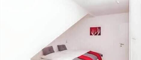 Habitació petita de color blanc amb teulada inclinada, un llit, una porta i un llum fixada a la paret.