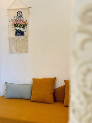 A bed with mustard-colored bedding and two pillows, one light blue and one mustard, against a white wall. A macramé wall hanging with a floral design is visible above the bed.