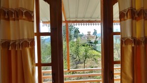 A view through a window with curtains, revealing a balcony with a railing and a building in the background. - Pears Garden Royal Suite (Kodaikanal)
