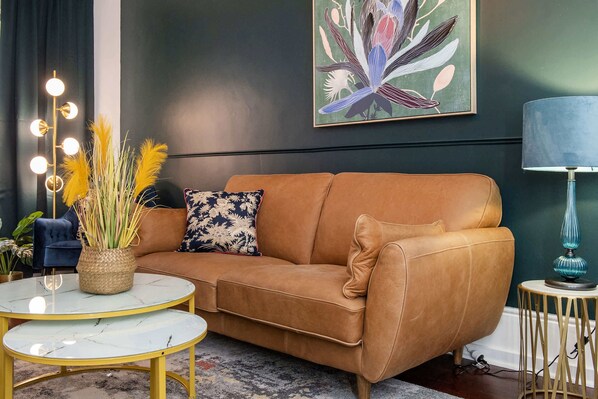 Ein Wohnzimmer mit braunem Ledersofa, einem runden weißen Tisch, einem blauen Sessel, einer Lampe und einem an der Wand hängenden Bild.