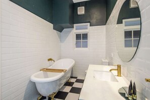 Ein Badezimmer mit weißen, rechteckigen Fliesen, einer freistehenden Badewanne mit goldfarbenen Armaturen, einem Waschbecken mit goldfarbenem Wasserhahn und einem Spiegel mit rundem Rahmen.
