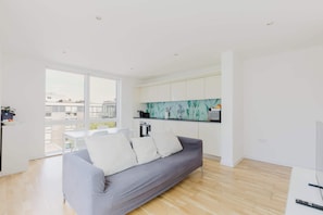A modern living room with a grey sofa, white cushions, a kitchen with white cabinets, and a large window.
